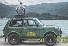 a man standing on top of a green car next to the water with mountains in the background