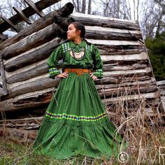 a woman wearing a green dress standing in front of a log cabin with her hands on her hips