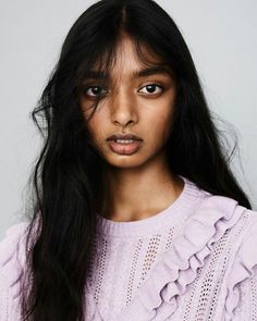 a woman with long black hair wearing a purple sweater and looking at the camera while standing in front of a gray background