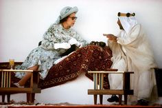 two women dressed in old fashioned clothing sitting on wooden chairs and talking to each other