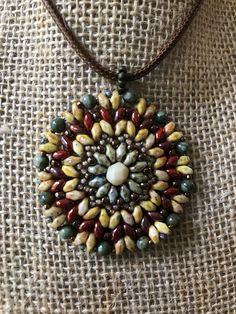 This colorful hand-beaded Mandala Necklace is made using many different style beads, and as you can see, very colorful!  It is warm colors, which include greens, browns, deep red, and brass colored beads. These include Super Duo beads, as well as assorted round beads in various sizes, and a large cut bead as the center piece. It is quite flexible, as shown in the one picture, but lays flat against your body. It measures 1-3/4 " inches in diameter, not including the beaded bale. It also includes Bohemian Round Brown Beads, Bohemian Brown Beaded Necklace, Bohemian Brown Beaded Round Necklaces, Handmade Brown Czech Glass Beads, Bohemian Brown Beaded Necklace With Czech Glass, Bohemian Brown Czech Glass Beaded Necklaces, Bohemian Brown Czech Glass Beaded Necklace, Earthy Multicolor Beaded Necklaces For Jewelry Making, Earthy Multicolor Beaded Necklace