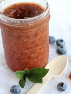 a jar filled with blueberries and sauce next to a spoon on a white tablecloth