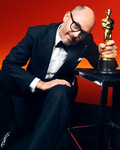 a man in a tuxedo leaning on a table with an oscar statue behind him