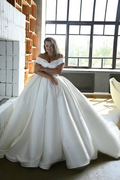 a woman in a white wedding dress leaning against a brick wall with her hands on her hips