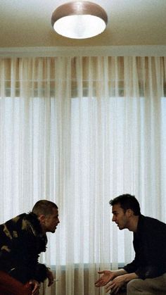 two men are talking in front of a window with sheer drapes on the windowsill