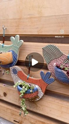 three colorful birds hanging from hooks on a wooden wall next to flowers and plants in vases