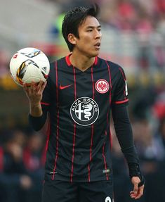 a man holding a soccer ball in his right hand and wearing a black uniform with red stripes