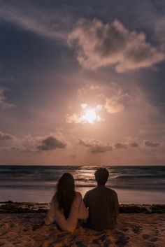 two people are sitting on the beach watching the sun go down over the water and clouds