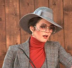 a woman wearing a gray hat and glasses talking to someone in front of a wooden wall