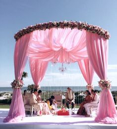 a group of people sitting under a pink canopy on top of a white floor next to the ocean