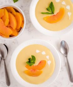 two white bowls filled with peaches on top of a marble counter next to spoons