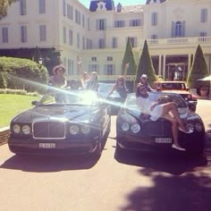 four women sitting on top of two cars in front of a mansion