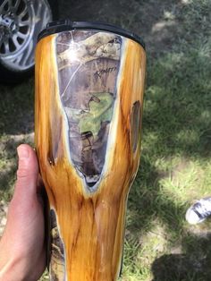 a person holding up a wooden cup in the grass with trees and mountains painted on it