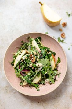 a pink bowl filled with green salad next to an apple on top of a table