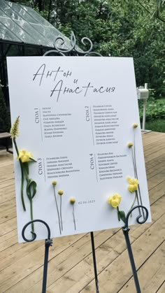 a sign with flowers on it sitting on top of a wooden deck in front of trees