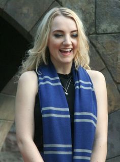 a woman with blonde hair wearing a blue and white striped scarf over her shoulders, standing in front of a stone wall