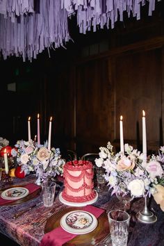 a table topped with a red cake covered in frosting next to tall candles and flowers