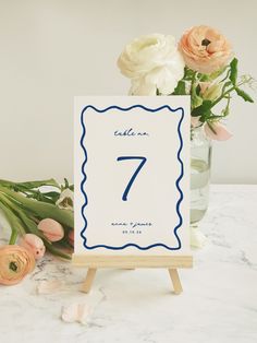 a table number with flowers in a vase on a marble countertop next to an easel