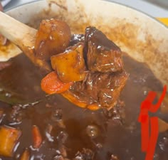 a wooden spoon full of beef and carrots in a pot with the lid open