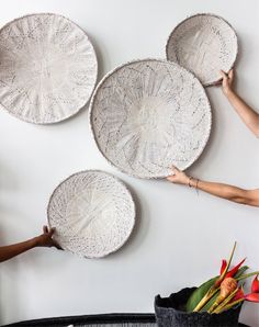 three white plates are hanging on the wall with flowers in front of them and two hands reaching for one