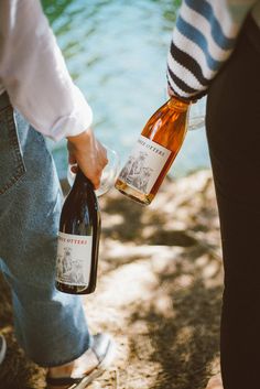 two people are holding wine bottles near the water