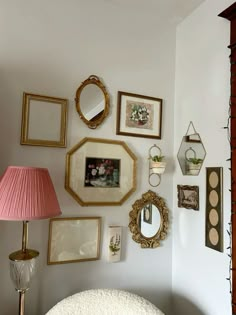 there are many framed pictures on the wall with a pink lamp next to it and a white chair