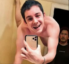 a shirtless man taking a selfie in front of his cell phone while another man looks on