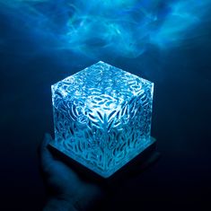 a hand holding a glowing cube in front of a blue background with swirling water behind it