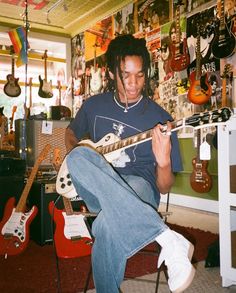 a young man sitting in a room playing an electric guitar with other guitars behind him