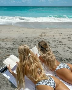 two women sitting on the beach reading a book