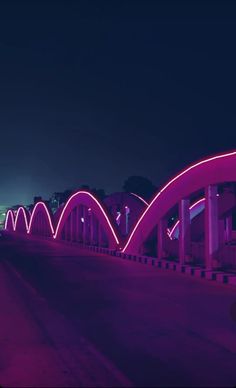 a bridge that is lit up at night with pink lights on the side and buildings in the background