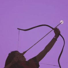 a woman holding an arrow in her right hand and looking up at the sky with purple skies behind her