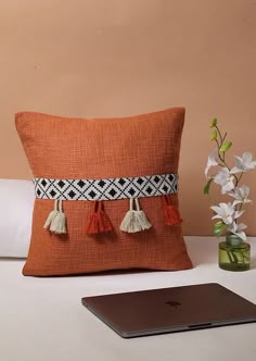 an orange pillow with tassels on it next to a laptop and flower vase