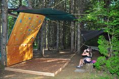 a woman sitting on a rock climbing platform in the woods next to a tent and trees