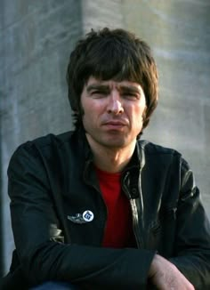 a young man with his arms crossed leaning on a railing looking at the camera while wearing a red shirt and black jacket