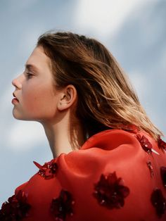 a woman with long hair wearing a red dress and looking off into the distance, against a cloudy blue sky