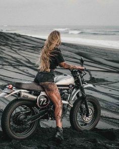 a woman sitting on top of a motorcycle next to the ocean with her legs crossed