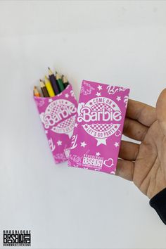 a hand holding a small pink box with colored pencils in it and the word barbie written on it