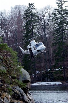 a helicopter is flying over the water near some trees and snow covered rocks in front of a body of water