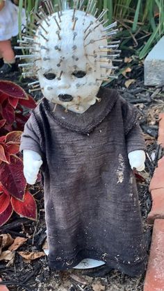 a creepy doll with spikes on its head is sitting in the ground next to some plants