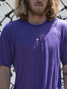 a man with long blonde hair wearing a purple shirt and silver necklace standing in front of graffiti covered wall