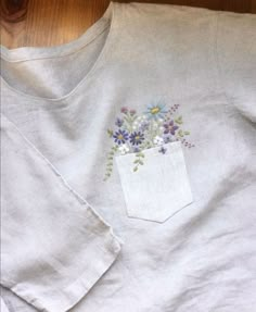 a white t - shirt with flowers embroidered on the front and back pocket, sitting on a wooden floor