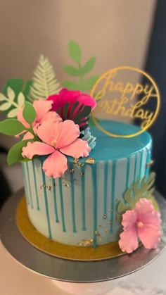 a blue and gold birthday cake with pink flowers on the top is sitting on a table