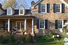 a stone house with black shutters and white flowers in the front yard, on a sunny day