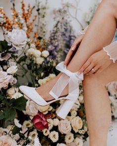 a woman's legs wearing white high heels with flowers in the back ground behind her