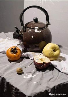 a tea kettle with apples and an orange on a white table cloth next to it