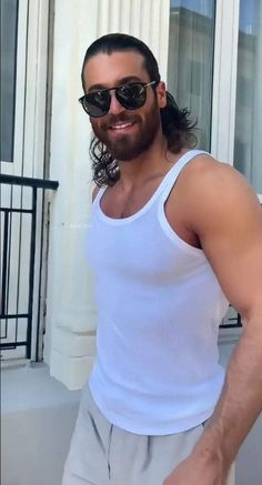 a man with long hair and sunglasses standing in front of a white building smiling at the camera