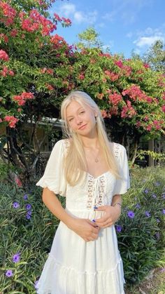 a woman in a white dress standing next to some pink and purple flowers on a sunny day