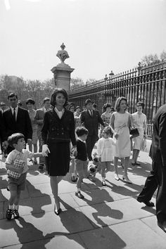 an old black and white photo of people walking on the sidewalk in front of a fence