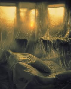 a bed covered in plastic sitting next to a window with the sun setting behind it